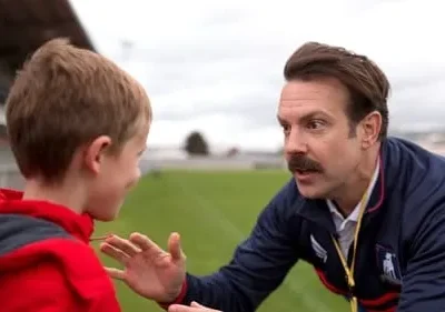 Um homem com bigode, de agasalho azul, fala com um menino de jaqueta vermelha em um campo. Ele gesticula de forma amigável.