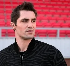 Homem em jaqueta estampada olhando pensativo para a direita, com arquibancadas vermelhas de estádio vazias ao fundo.