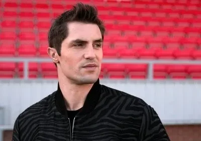 Homem em jaqueta estampada olhando pensativo para a direita, com arquibancadas vermelhas de estádio vazias ao fundo.