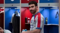 Homem barbudo com camisa esportiva olha sério na frente de armários vermelhos e azuis de vestiário.