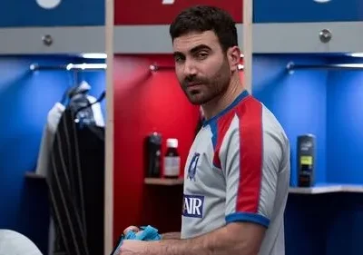 Homem barbudo com camisa esportiva olha sério na frente de armários vermelhos e azuis de vestiário.