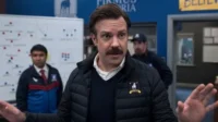 Um treinador com bigode e jaqueta puffer gesticula. Dois assistentes atrás e um quadro tático. Clima esportivo enérgico.