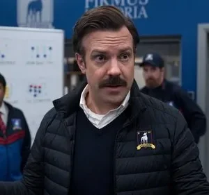 Um treinador com bigode e jaqueta puffer gesticula. Dois assistentes atrás e um quadro tático. Clima esportivo enérgico.