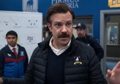 Um treinador com bigode e jaqueta puffer gesticula. Dois assistentes atrás e um quadro tático. Clima esportivo enérgico.