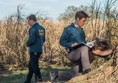 Dois investigadores em um campo com capim alto. Um ajoelha examinando um veado com chifres, anotando. Clima sombrio e rural.