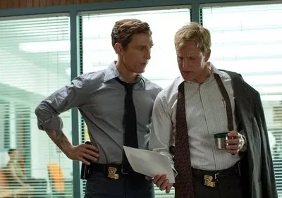 Dois homens sérios em um escritório. Um loiro com blazer e papéis, o outro de camisa cinza. Clima de investigação.