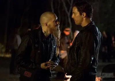 Dois homens sérios, um com barba longa, o outro de cabelo escuro, se encaram. Vestem jaquetas de couro à noite, com figuras no fundo. Clima tenso.