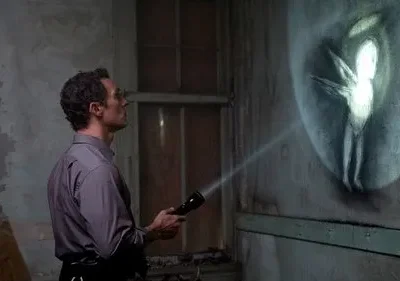 Homem com lanterna ilumina figura alada fantasmagórica brilhando na parede descascada de um quarto escuro e abandonado. Misterioso.