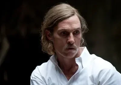 Homem de cabelo loiro e bigode, camisa branca, com olhar intenso em fundo escuro e misterioso. Clima sombrio.