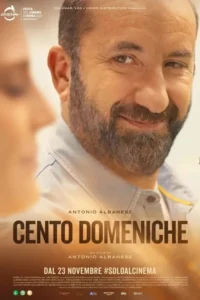 Um homem barbudo sorri calorosamente para a câmera,com uma mulher desfocada à esquerda. Fundo suave e iluminado.