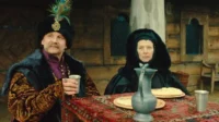 Um homem elegante com chapéu de penas e uma mulher de capuz escuro sentam-se à mesa em cenário rústico. Trajes de época.