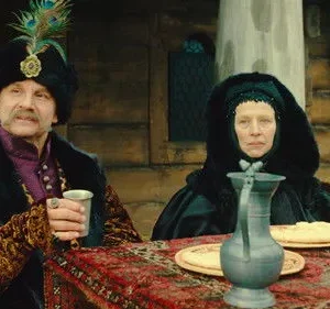 Um homem elegante com chapéu de penas e uma mulher de capuz escuro sentam-se à mesa em cenário rústico. Trajes de época.