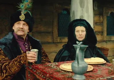 Um homem elegante com chapéu de penas e uma mulher de capuz escuro sentam-se à mesa em cenário rústico. Trajes de época.