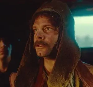 Um homem com barba e capuz, rosto sujo, olha apreensivo. Atrás, um jovem observa na penumbra em cenário rústico.