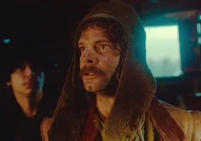 Um homem com barba e capuz, rosto sujo, olha apreensivo. Atrás, um jovem observa na penumbra em cenário rústico.