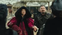 Mulher de casaco vermelho e homem barbudo com túnica bordada sorriem,de mãos dadas,em um vilarejo. Clima festivo.