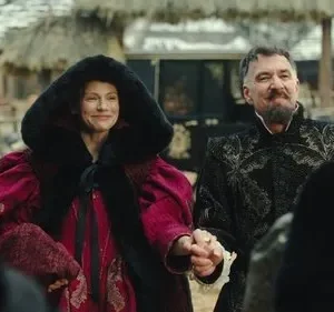 Mulher de casaco vermelho e homem barbudo com túnica bordada sorriem, de mãos dadas, em um vilarejo. Clima festivo.