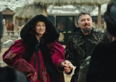 Mulher de casaco vermelho e homem barbudo com túnica bordada sorriem, de mãos dadas, em um vilarejo. Clima festivo.