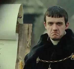 Homem jovem com franja, casaco preto e expressão carrancuda, perto de um pergaminho antigo. Cenário histórico.