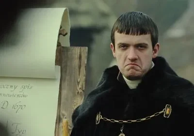 Homem jovem com franja, casaco preto e expressão carrancuda, perto de um pergaminho antigo. Cenário histórico.