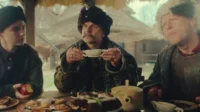 Três pessoas com trajes históricos e chapéus em uma mesa rústica cheia de comida. Expressões sérias em um ambiente campestre.