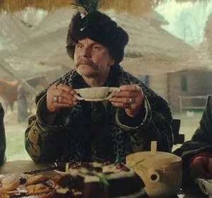 Três pessoas com trajes históricos e chapéus em uma mesa rústica cheia de comida. Expressões sérias em um ambiente campestre.