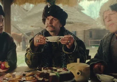 Três pessoas com trajes históricos e chapéus em uma mesa rústica cheia de comida. Expressões sérias em um ambiente campestre.