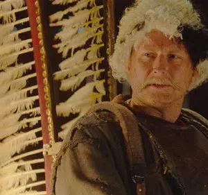Homem com bigode e cabelo metade branco, metade preto, veste roupas rústicas. Grande estrutura de penas brancas atrás dele.