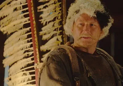 Homem com bigode e cabelo metade branco, metade preto, veste roupas rústicas. Grande estrutura de penas brancas atrás dele.