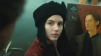 Jovem em chapéu preto e roupa estampada olha atentamente. Atrás dela,um cavalete com um retrato feminino. Clima histórico.