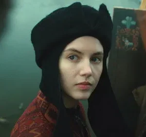 Jovem em chapéu preto e roupa estampada olha atentamente. Atrás dela, um cavalete com um retrato feminino. Clima histórico.