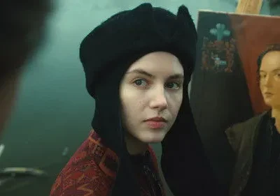 Jovem em chapéu preto e roupa estampada olha atentamente. Atrás dela, um cavalete com um retrato feminino. Clima histórico.