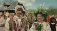Jovem casal em trajes tradicionais sorrindo. Ela usa uma coroa de flores e segura um buquê;ele,um chapéu. Multidão rural ao fundo.