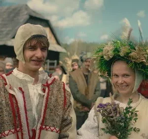Jovem casal em trajes tradicionais sorrindo. Ela usa uma coroa de flores e segura um buquê; ele, um chapéu. Multidão rural ao fundo.