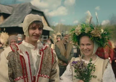 Jovem casal em trajes tradicionais sorrindo. Ela usa uma coroa de flores e segura um buquê; ele, um chapéu. Multidão rural ao fundo.
