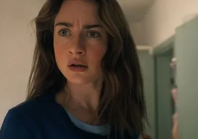 Jovem mulher com cabelos castanhos e olhos arregalados, boca entreaberta, expressa choque em corredor. Clima tenso.