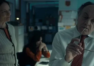 Homem pensativo com óculos na boca, mulher atenta e pessoa cansada em escritório escuro. Atmosfera séria e dramática.