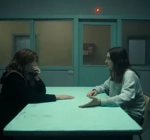 Duas mulheres em sala escura e fria. Uma pensativa, outra gesticula. Homem observa ao fundo. Clima tenso e misterioso.