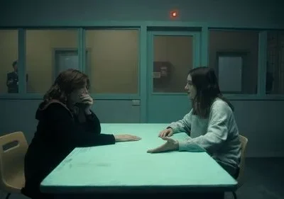 Duas mulheres em sala escura e fria. Uma pensativa, outra gesticula. Homem observa ao fundo. Clima tenso e misterioso.