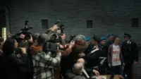 Fotógrafos cercam um homem e policiais escoltam uma mulher em um tribunal. Clima tenso e caótico com intensa atenção da mídia.