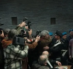 Fotógrafos cercam um homem e policiais escoltam uma mulher em um tribunal. Clima tenso e caótico com intensa atenção da mídia.