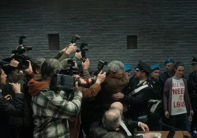 Fotógrafos cercam um homem e policiais escoltam uma mulher em um tribunal. Clima tenso e caótico com intensa atenção da mídia.