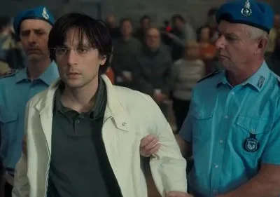 Jovem de óculos e jaqueta clara é escoltado por dois oficiais de farda azul em tribunal, com público ao fundo. Clima sério.