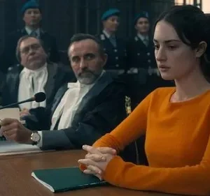 Mulher séria de suéter laranja sentada em tribunal. Juízes e policiais ao fundo. Clima de drama legal e tensão.