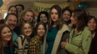 Mulher séria no centro, rodeada por um grupo sorridente de diversas idades em evento com balões e faixa de boas-vindas ao fundo.