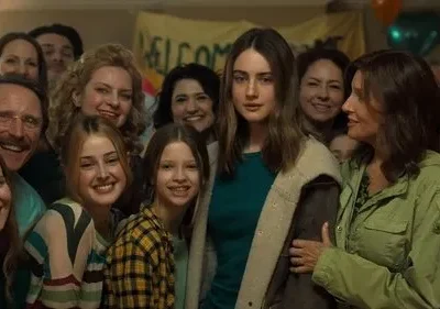 Mulher séria no centro, rodeada por um grupo sorridente de diversas idades em evento com balões e faixa de boas-vindas ao fundo.