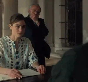 Mulher sentada com expressão séria em frente a papéis, um homem mais velho observa por trás. Ambiente formal com colunas, clima tenso.