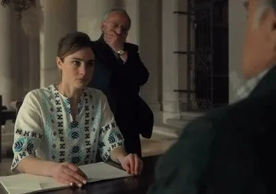 Mulher sentada com expressão séria em frente a papéis, um homem mais velho observa por trás. Ambiente formal com colunas, clima tenso.