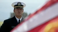 Homem sério em uniforme da marinha, com boné branco, olhando fixamente. Bandeira americana borrada no primeiro plano.