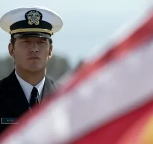 Homem sério em uniforme da marinha, com boné branco, olhando fixamente. Bandeira americana borrada no primeiro plano.